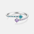A silver-toned open ring band featuring two round gemstones, one turquoise blue and one purple, with small clear accent stones along the band, shown on a light gray background.