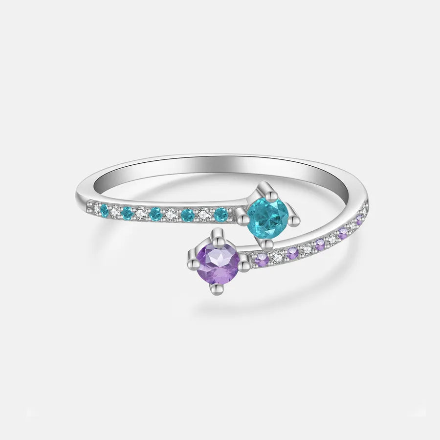 A silver-toned open ring band featuring two round gemstones, one turquoise blue and one purple, with small clear accent stones along the band, shown on a light gray background.