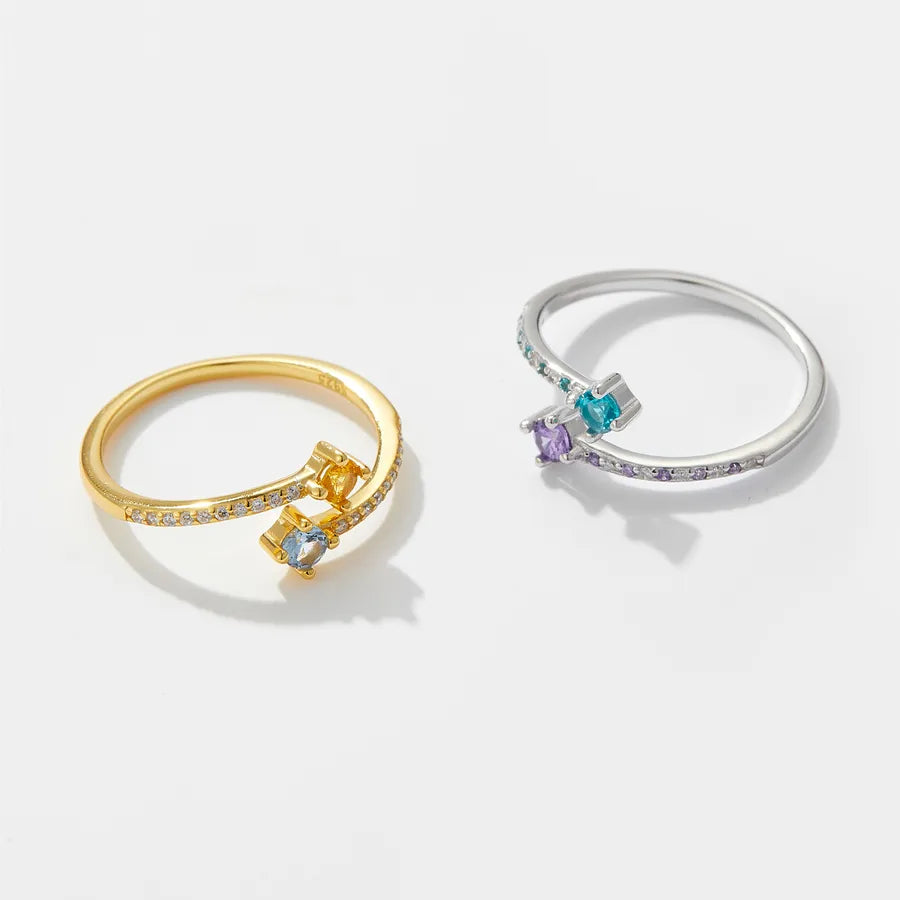 Two minimalist open-band rings on a clean white background, one in yellow gold with a small blue gemstone and tiny clear stones, and one in silver with a small purple gemstone and tiny clear stones.