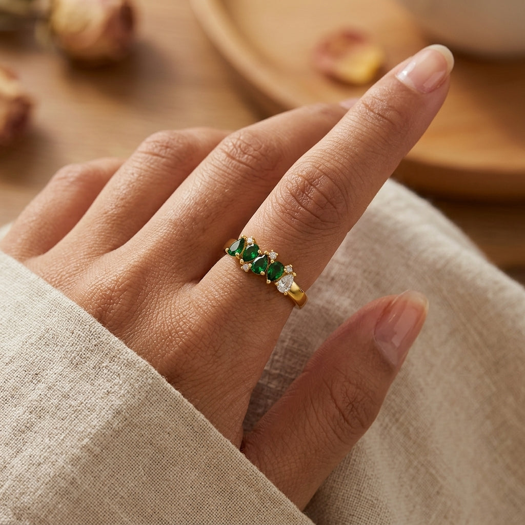 A close-up of a hand wearing a gold ring adorned with three emerald-cut green gemstones and small clear accent stones, set against a softly blurred warm-toned background.