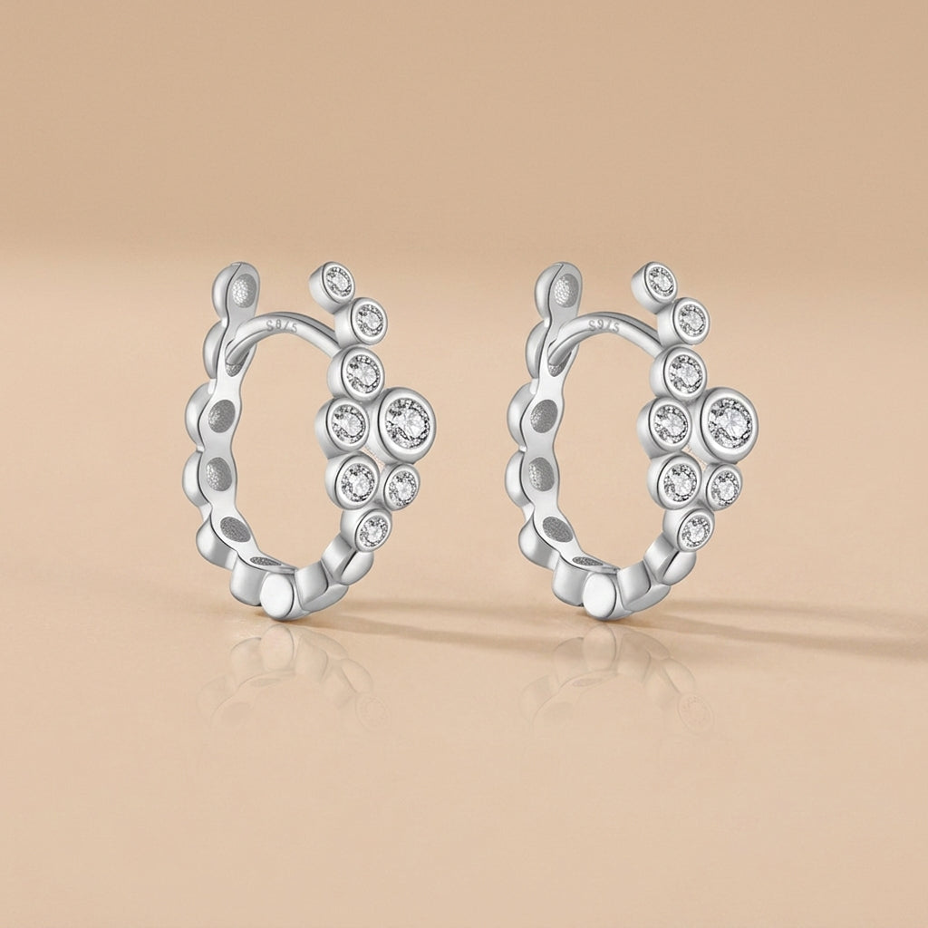 A pair of silver crescent-shaped hoop earrings adorned with clear round-cut gemstones, displayed on a warm beige background.