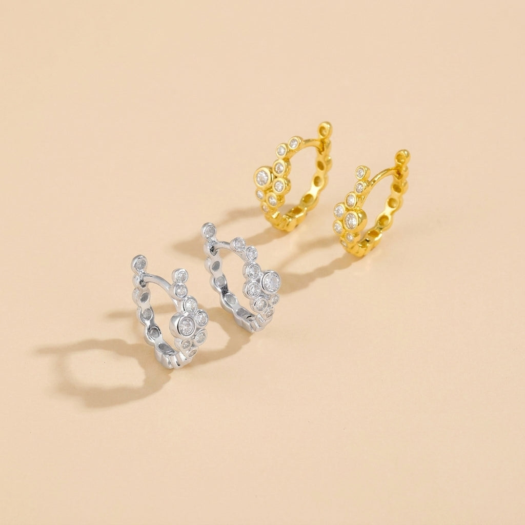 Two small hoop earrings on a warm beige background, one in yellow gold and one in silver, both decorated with clear crystal-like stones.